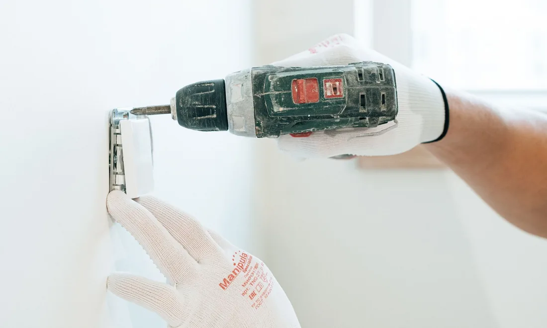 Hände in Arbeitshandschuhen montieren mit einem Akkuschrauber eine Steckdose an einer Wand